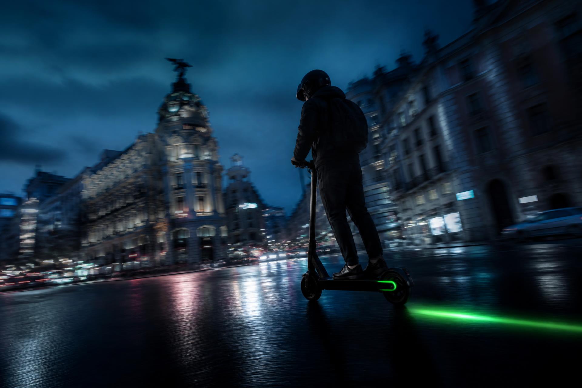 Persona circulando en patinete eléctrico por una avenida urbana al anochecer, con estelas de luz sobre el asfalto mojado